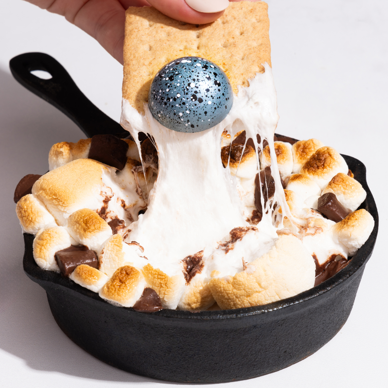A close-up of a blue S'mores chocolate getting scooped up out of a black cast iron skillet featuring chocolate and toasted marshmallows. The S'mores chocolate is being scooped up out of the skilled by a graham cracker. 